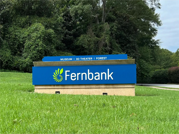 Fernbank Museum sign with a blue background and Fernbank logo, which is green and white. The background is a lush amount of greenery.